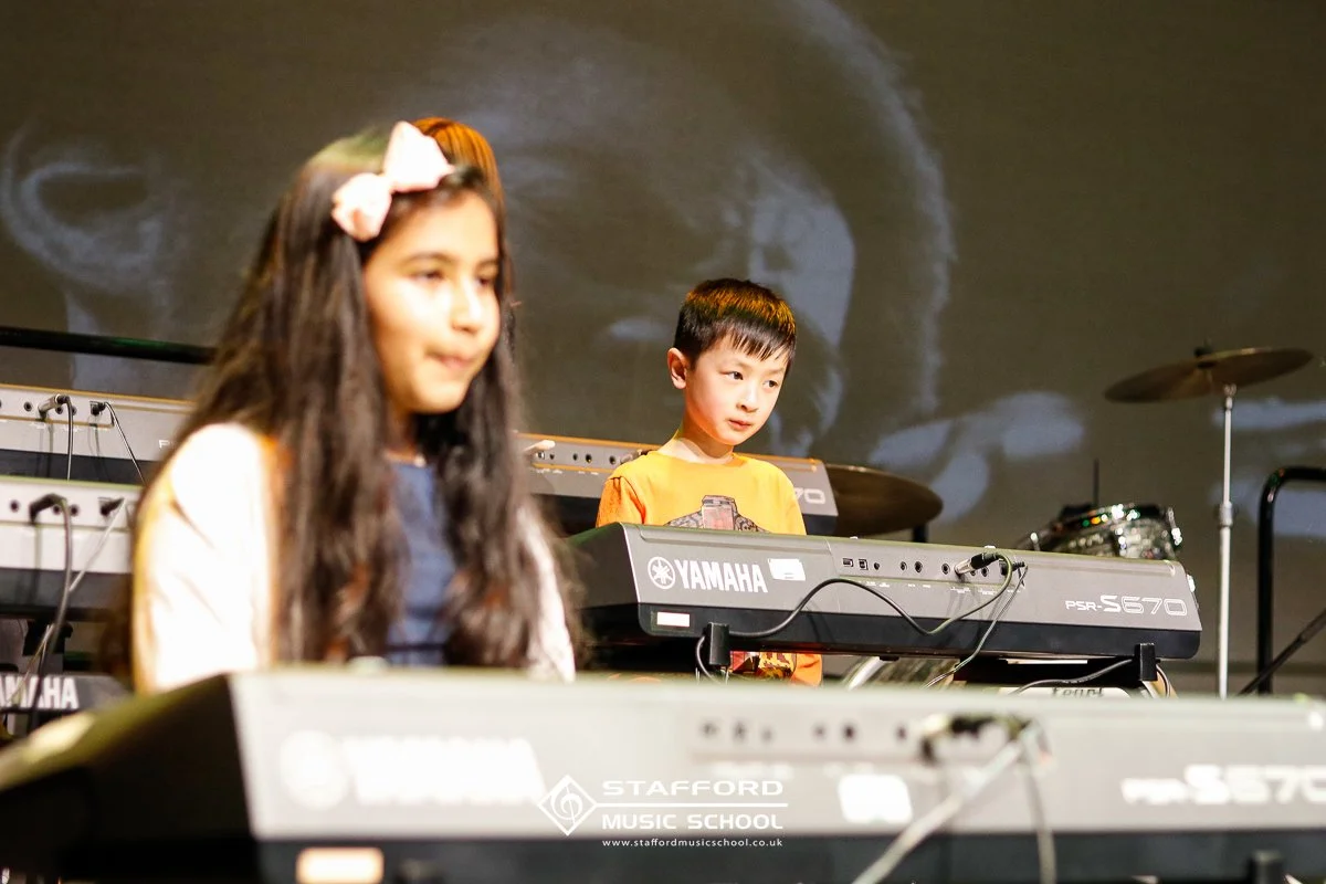 Two children, a girl with long dark hair and a boy with short dark hair, are playing Yamaha keyboards during a music class. The girl is wearing a pink headband with a bow and a yellow cardigan, while the boy is in a yellow shirt. There is a drum set 