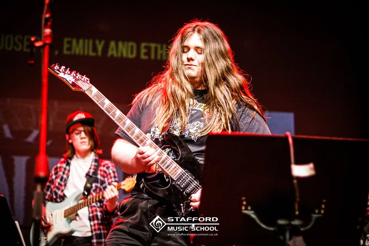 Two young women playing electric guitars on stage during a performance, with a personalized logo of Stafford Music School and a background projection with text partially visible.