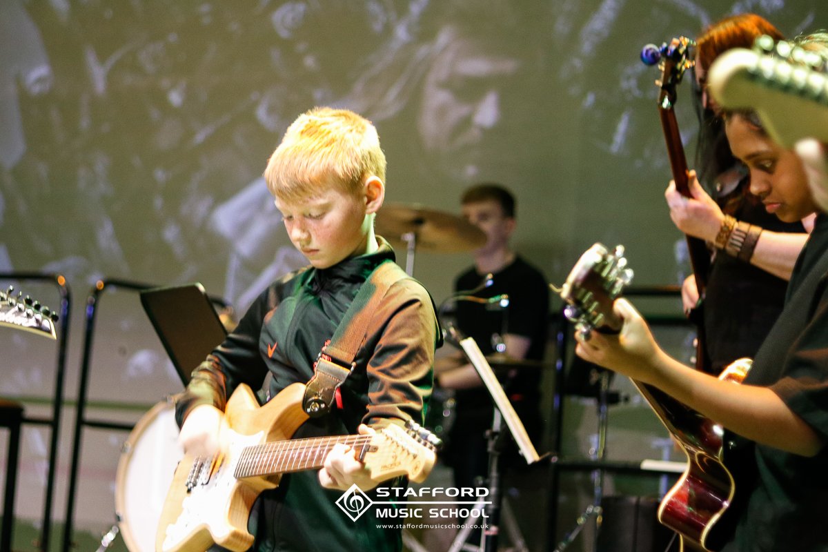 Young boy with red hair playing an electric guitar on stage, with two other guitarists and a drummer in the background during a band performance at Stafford Music School.