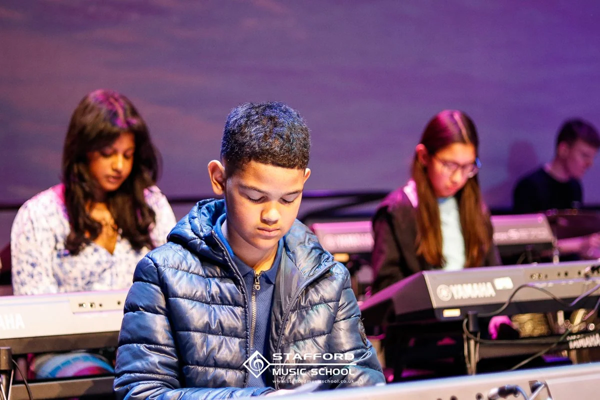 Children playing keyboards on stage during a music class at Stafford Music School
