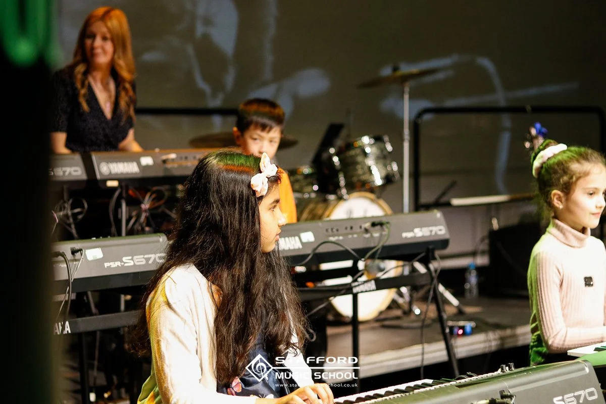 Children performing on stage with musical instruments, including keyboards and drums, with a woman in the background.