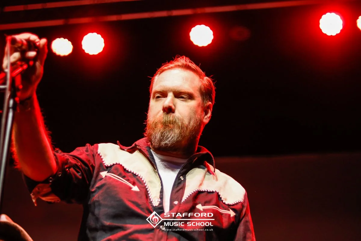 A man with a beard and mustache on stage holding a microphone stand, with red stage lights overhead. The logo of Stafford Music School is visible at the bottom of the image.