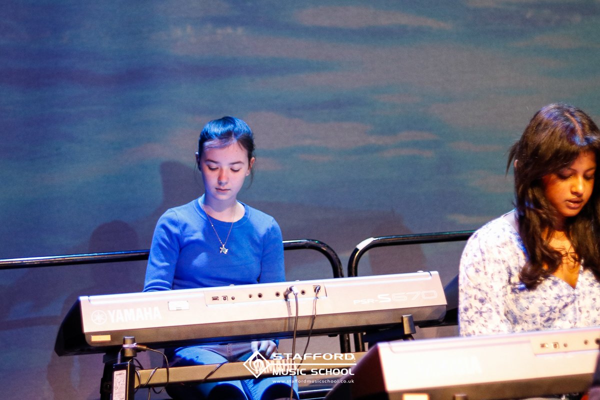 Two young girls playing keyboards during a performance at Stafford Music School, with a dark background.