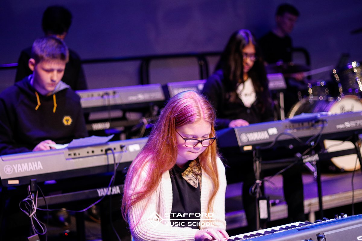 Music students performing on Yamaha keyboards during a concert at Tafford Music School, with a young woman with long red hair and glasses in the foreground and other students in the background.