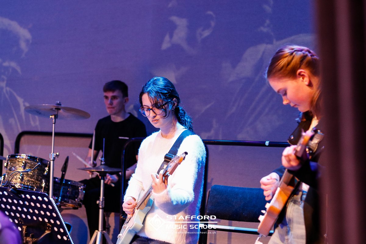 Three young musicians performing on stage: a drummer in the background, a girl with glasses playing an electric guitar in the center, and a girl playing an acoustic guitar on the right.