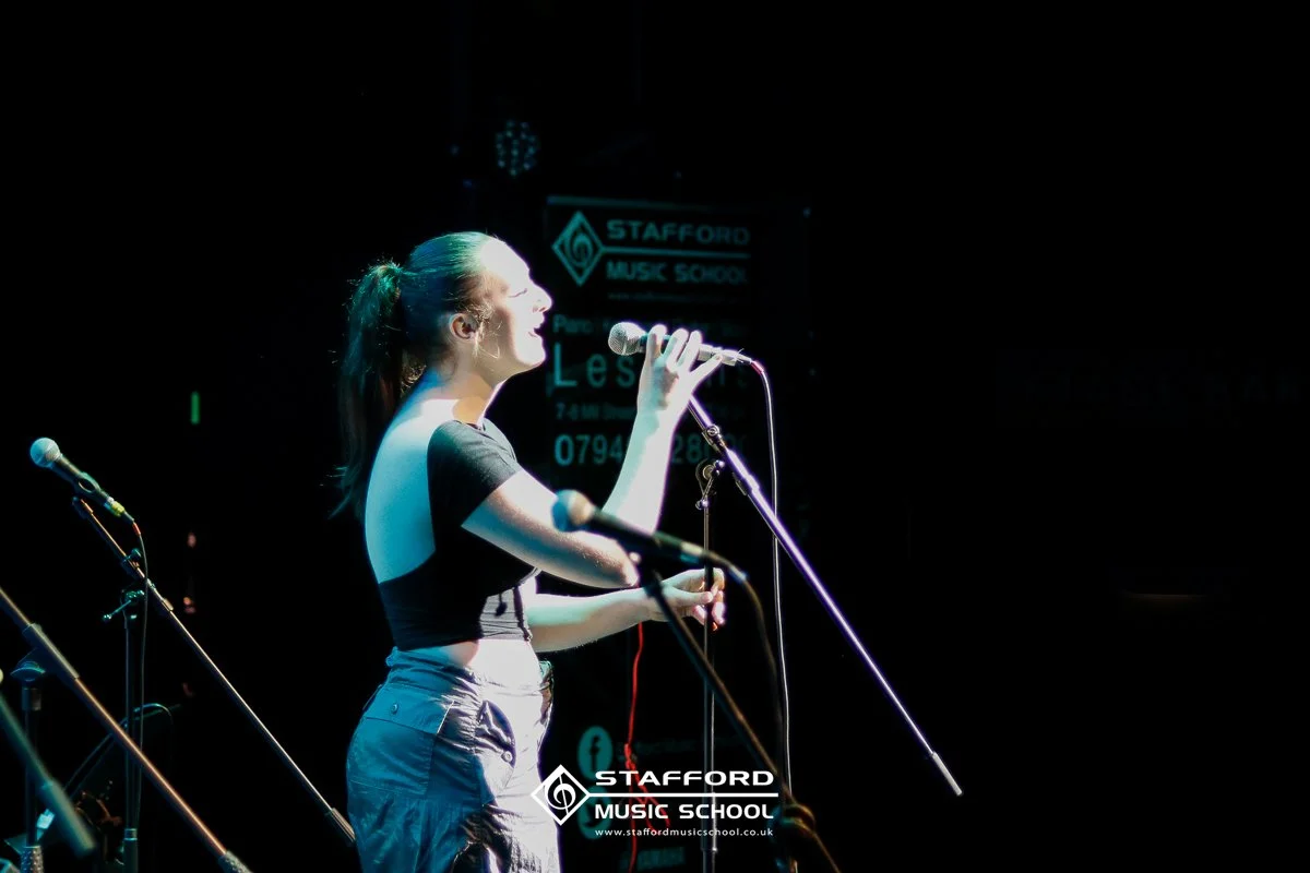 Young woman singing into a microphone on stage at Stafford Music School, with stage lights highlighting her and a dark background.