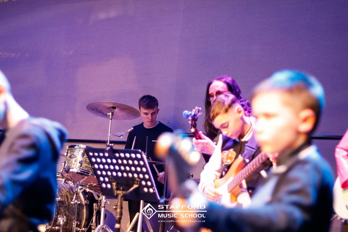 Children playing musical instruments during a performance at Stafford Music School, with a person playing drums, a girl with glasses on guitar, and other children playing guitars, on a stage with purple lighting.