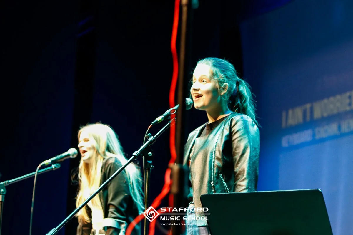 Two female singers performing on stage with microphones, one in focus and the other slightly blurred, at Stafford Music School.