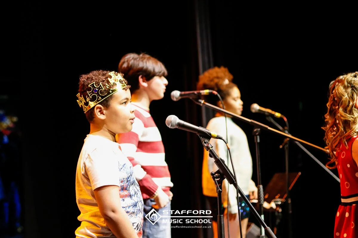Children performing on stage, standing in front of microphones at Stafford Music School.