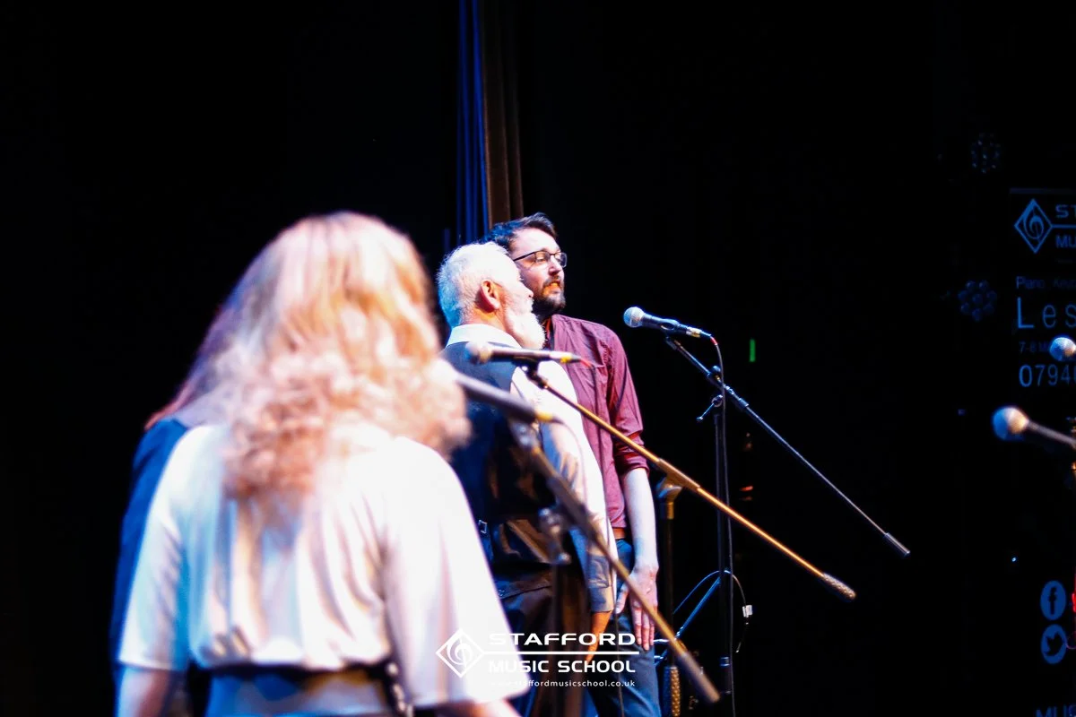 Three musicians on stage at Stafford Music School, with microphones set up, dark background, and stage lighting highlighting the performers.