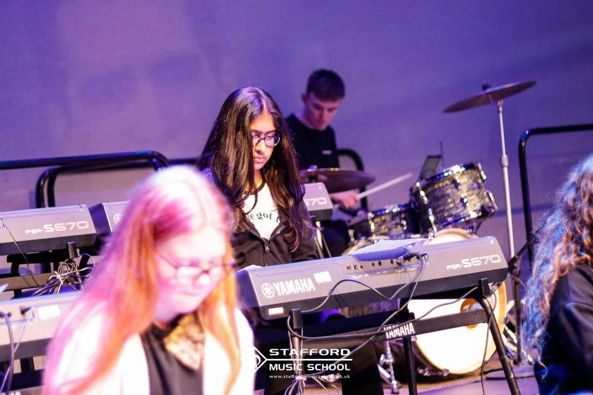 A young woman with long brown hair, glasses, and a white shirt is playing a Yamaha keyboard at a music performance, with a drummer in the background.