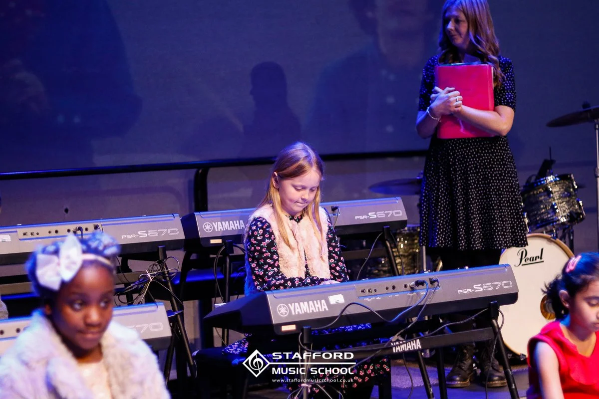 Young girl playing a keyboard on stage during a school music performance with other children and a woman holding a pink folder in the background.