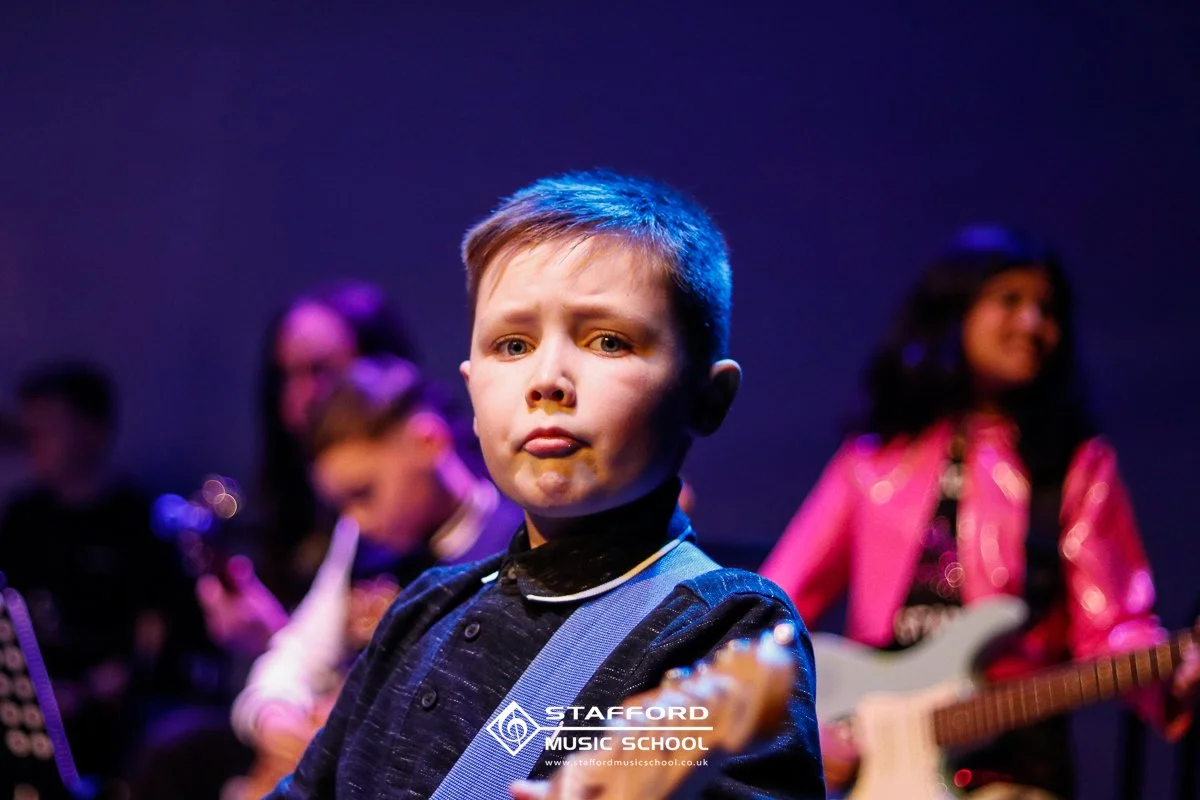 Young boy with a serious expression standing on stage, playing an electric guitar, with two women in the background, one playing an electric guitar and the other possibly singing or speaking, all at Stafford Music School.