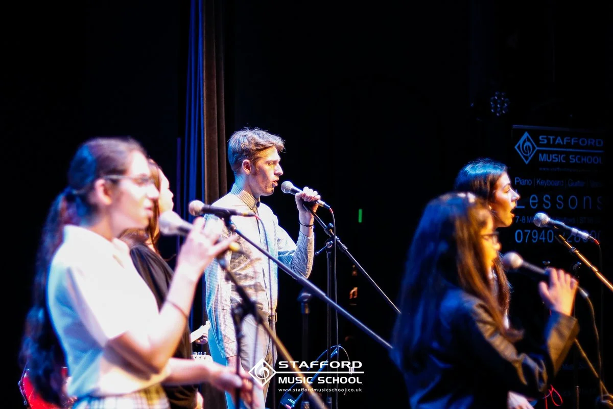 Group of young people singing into microphones on stage at Stafford Music School.