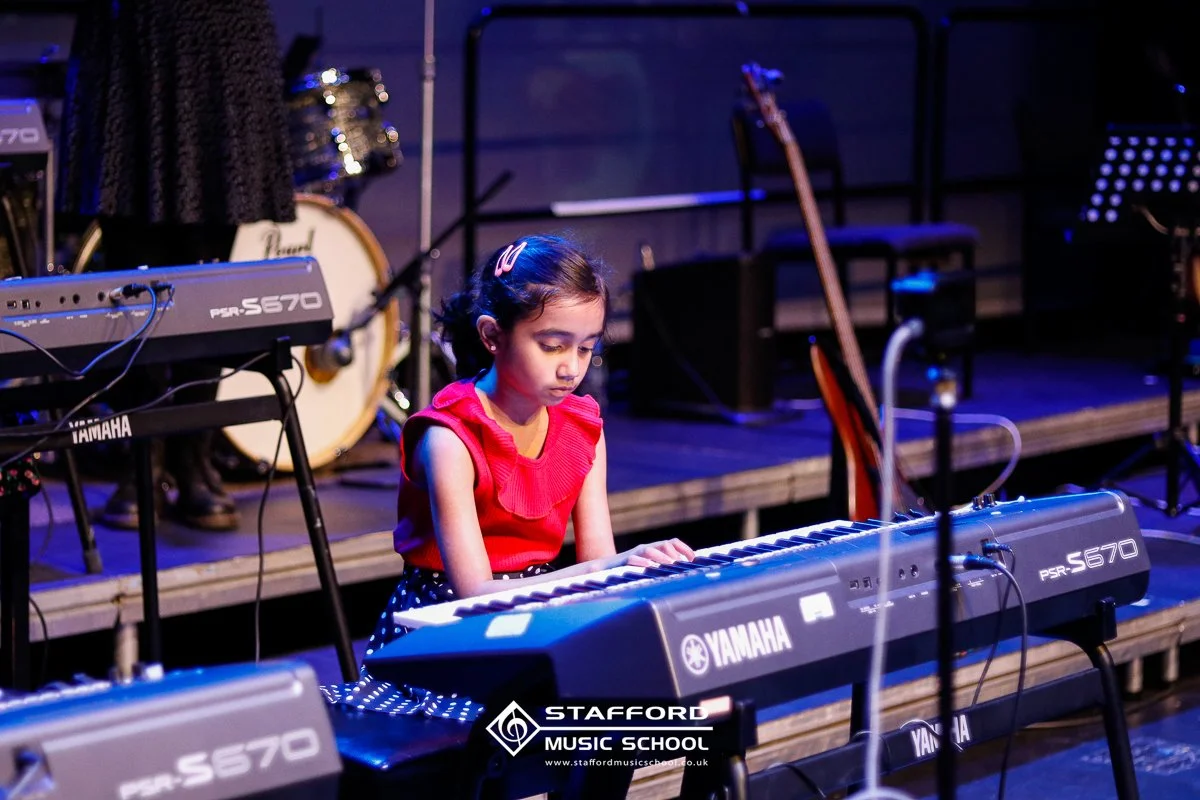 A young girl in a red top playing a Yamaha keyboard on stage during a music performance.