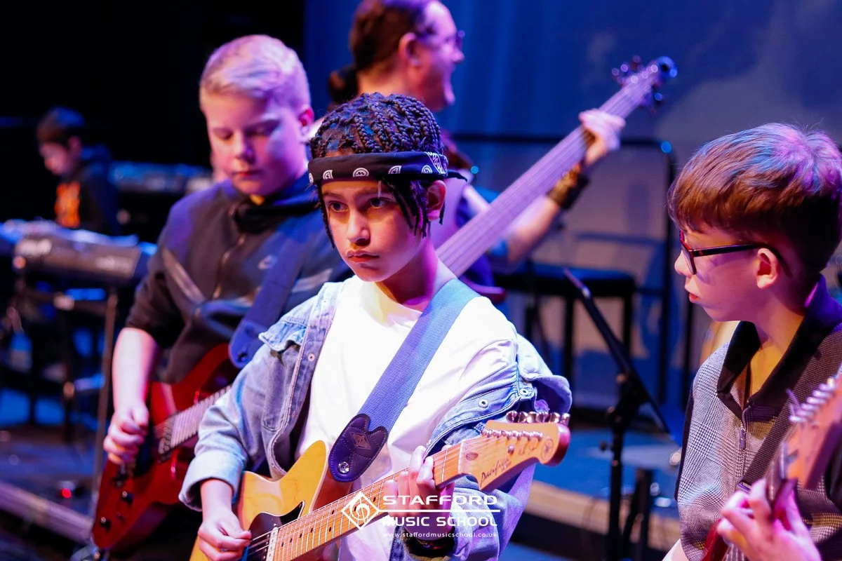 Young students practicing guitar and keyboard on stage in a music class at Stafford Music School.