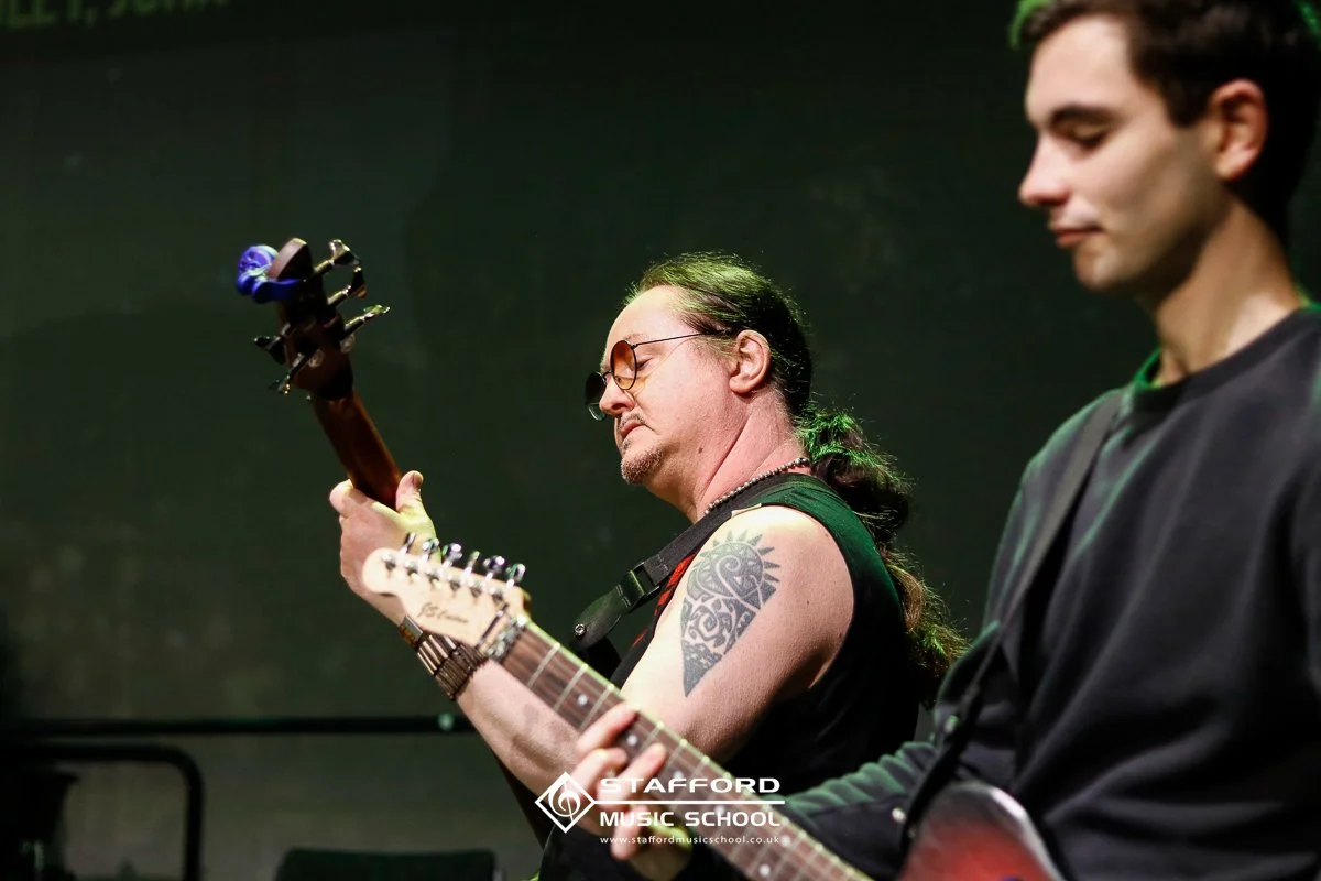Two musicians playing guitars in a dimly lit room, with the Stafford Music School logo in the bottom center.