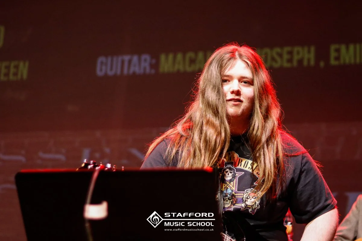 Young person with long brown hair sitting at a keyboard on stage, with a dark background and yellow and purple text projected on the wall behind them.