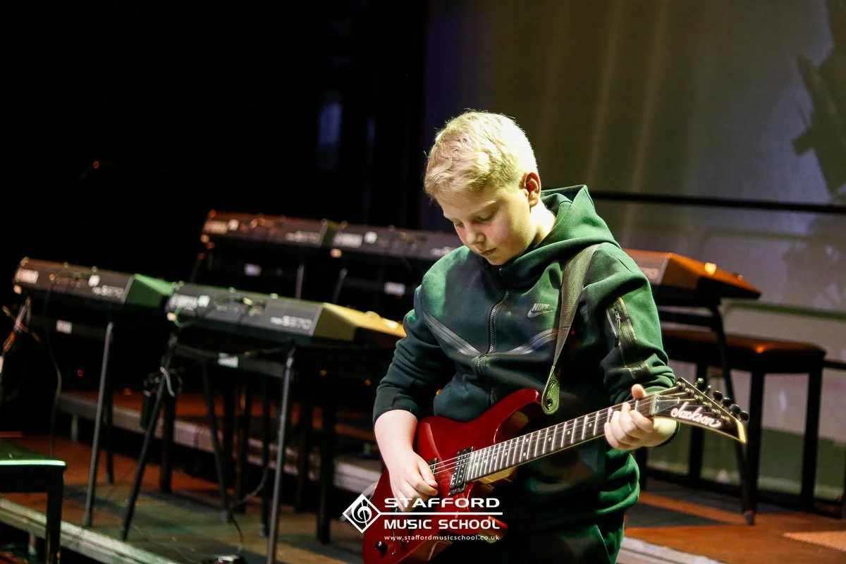 A young boy playing a red electric guitar on stage at Stafford Music School, with various keyboards and musical equipment in the background.