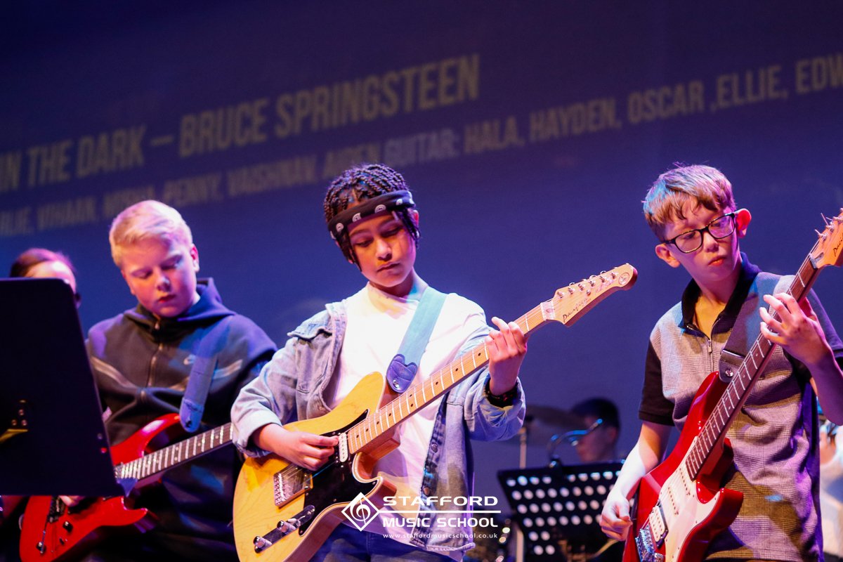 School children performing on stage with electric guitars at Stafford Music School, with a blue background and a partially visible projection of band members' names.