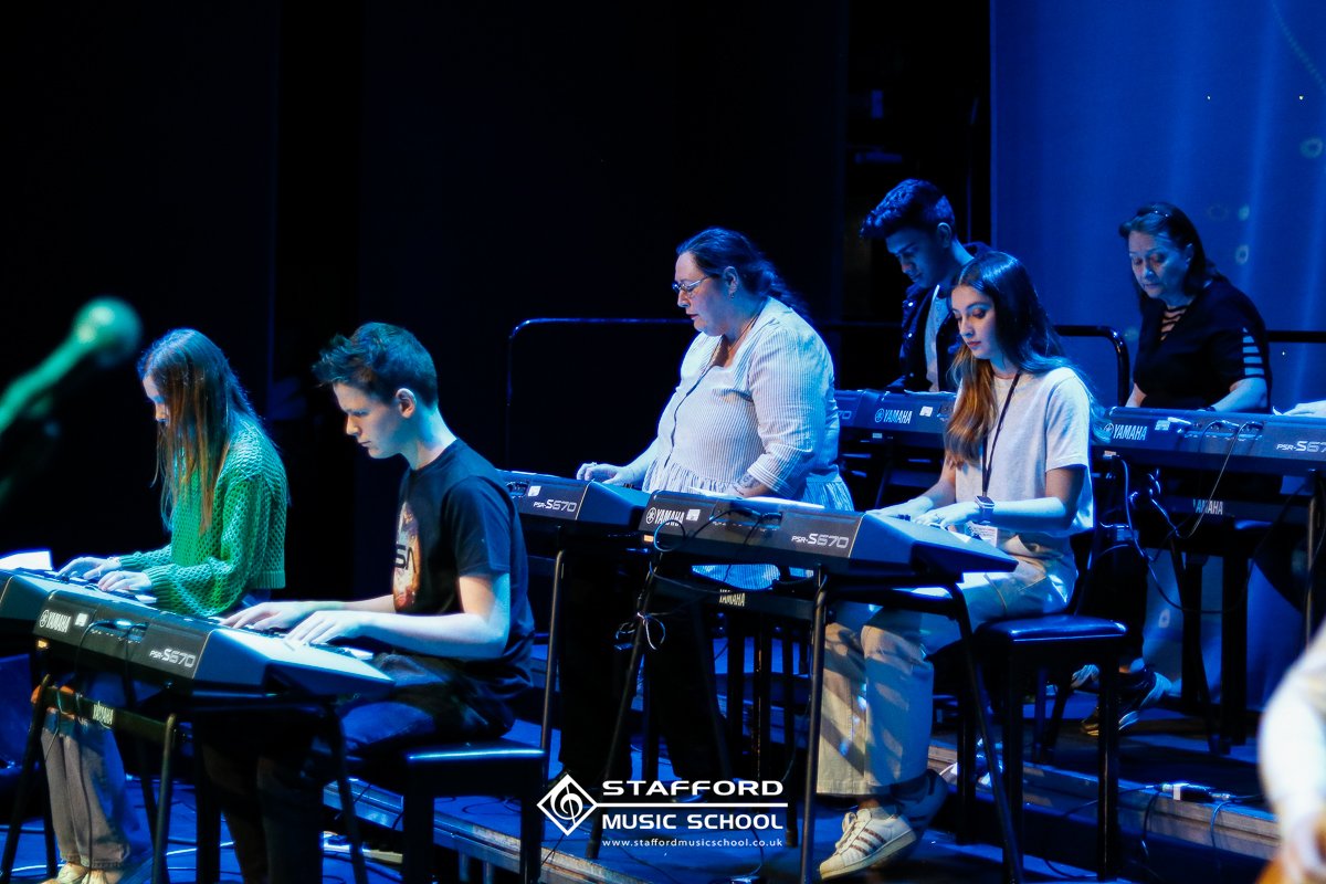 Group of people playing Yamaha synthesizers on stage during a music rehearsal at Stafford Music School.
