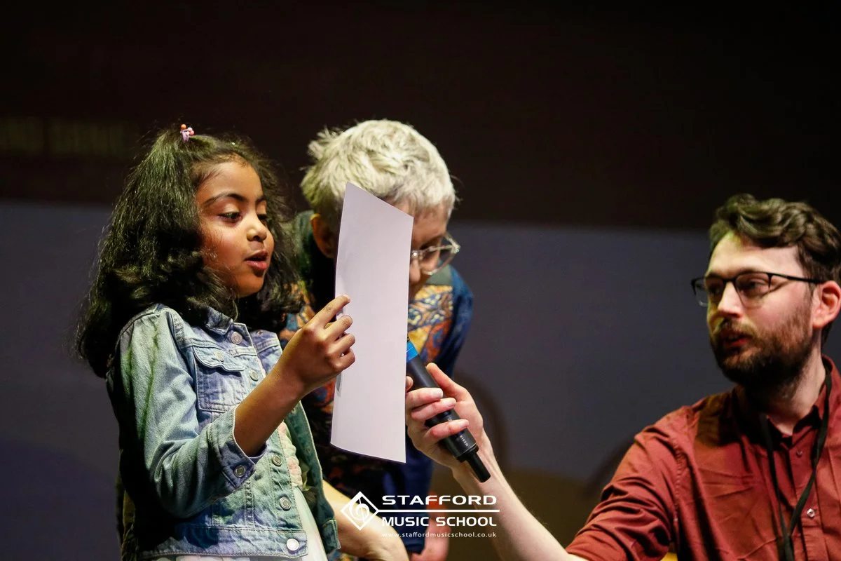 A young girl with dark curly hair holding a piece of paper and speaking into a microphone. An older woman and a man with glasses are listening attentively.