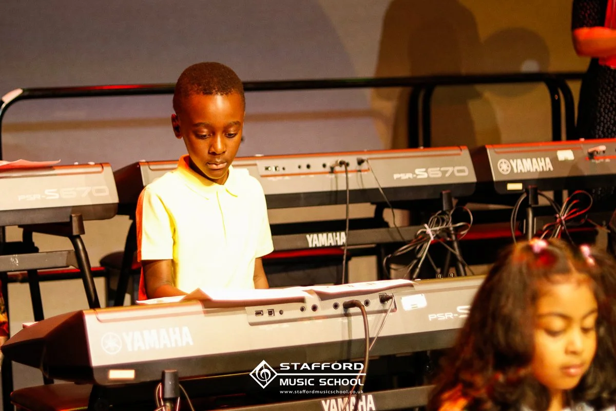 A young boy playing a Yamaha keyboard at Stafford Music School with other children around.