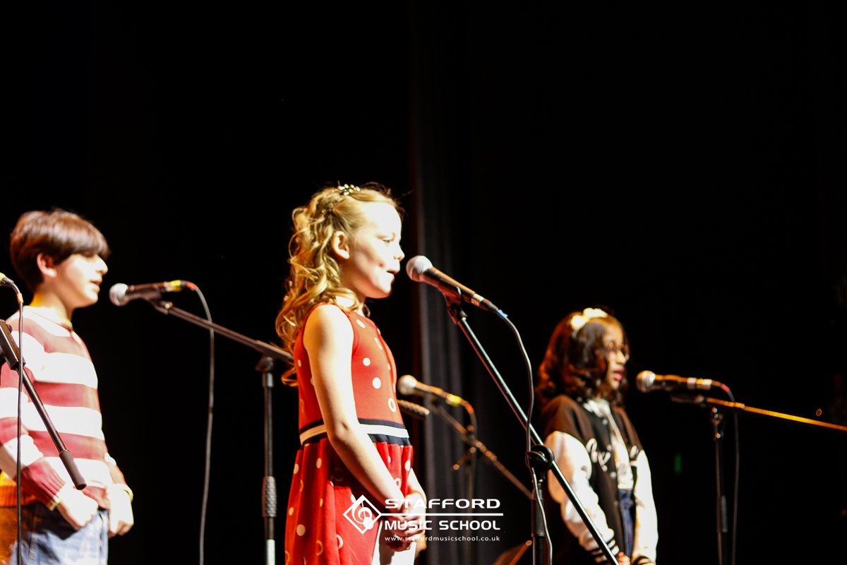 Three young girls standing at microphones on stage during a performance at Stafford Music School, with dark background lights focusing on them.
