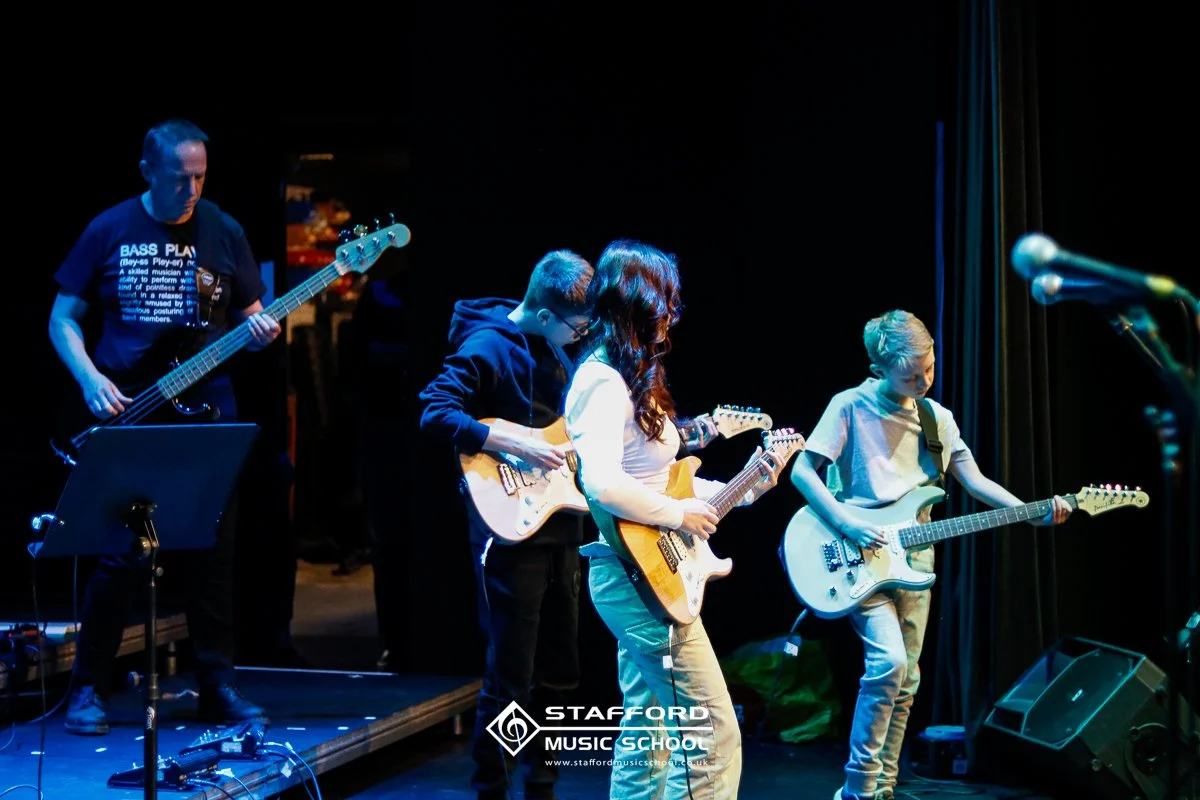 A group of five musicians on stage, playing electric guitars and bass, with a dark background and stage lighting.