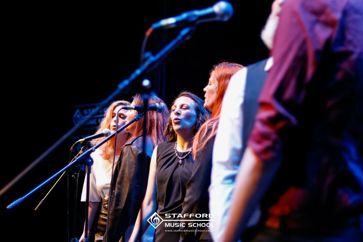 Group of women singing on stage with microphones at Stafford Music School.