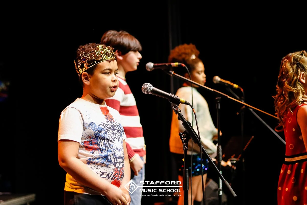Children performing on stage at Stafford Music School, with microphones and stage lighting, one wearing a crown and a T-shirt with a lion design.