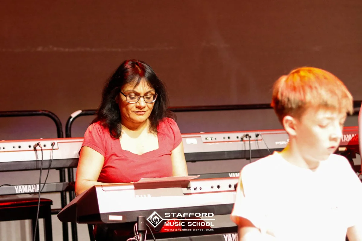 A woman with dark hair and glasses playing a keyboard on stage, with music equipment in the background, during a music class at Stafford Music School.