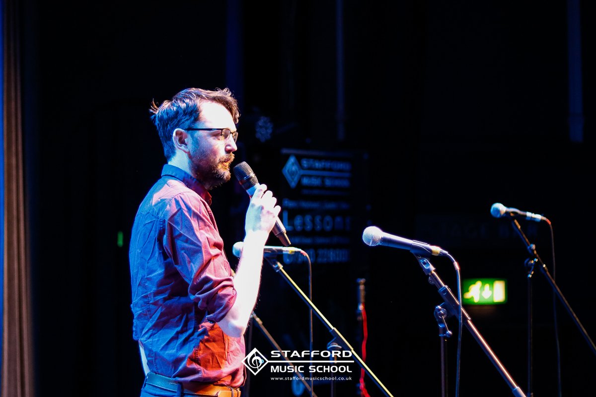 A man with glasses and a beard is holding a microphone on stage, with two additional microphones on stands in front of him. He appears to be preparing to speak or perform. The background includes signage for Stafford Music School and an emergency exi
