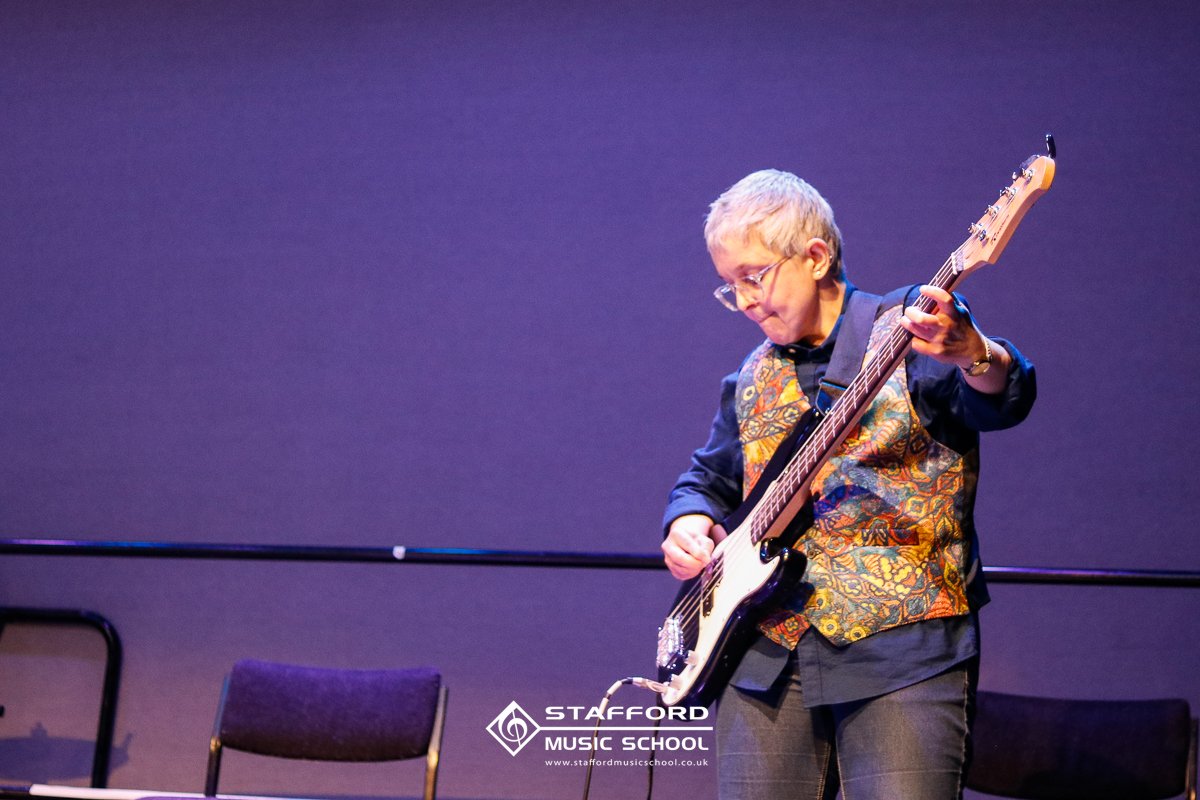 A person with short gray hair and glasses playing an electric guitar on stage. They are wearing a black shirt and a colorful vest, standing in front of a purple background with chairs and a logo for Stafford Music School.