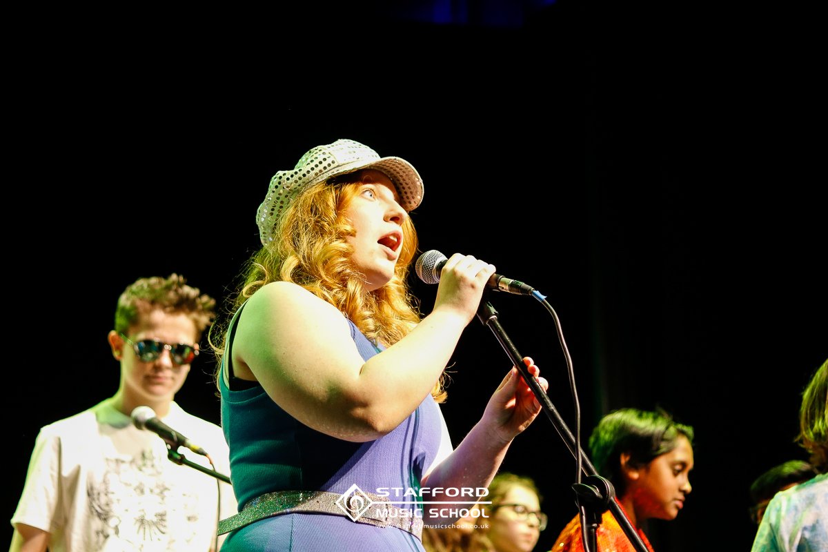 A group of young women performing on stage, with one girl singing into a microphone and wearing a white hat, and others in the background with microphones and colorful outfits.