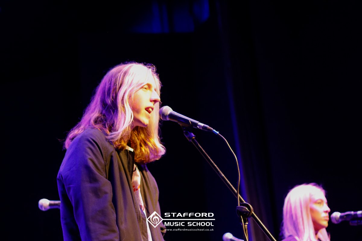 A young woman with long, wavy blonde hair and glasses singing into a microphone on stage with blue and purple lighting, with another woman in the background also at a microphone, at Stafford Music School.