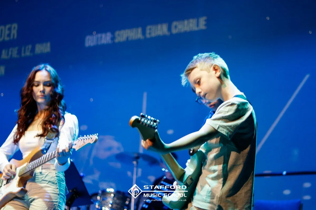 Two young children playing electric guitars on stage during a music performance at Stafford Music School.