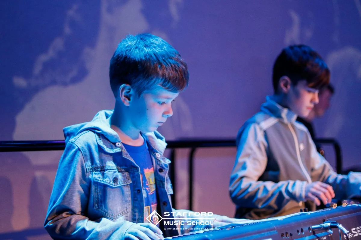 Two young boys are playing electronic keyboard instruments, possibly synthesizers, in a dimly lit room, with a blue and purple background. The logo of Stafford Music School is visible at the bottom.