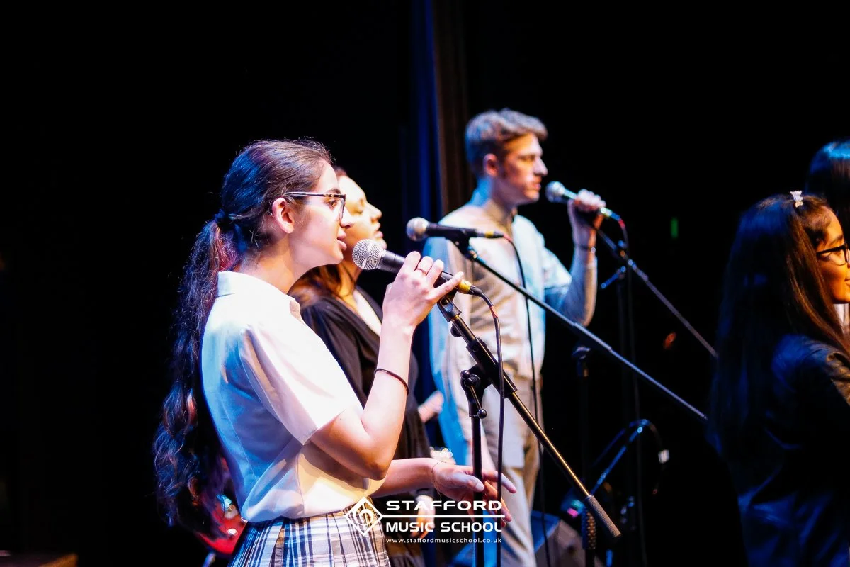 Group of young people singing into microphones on stage at Stafford Music School.