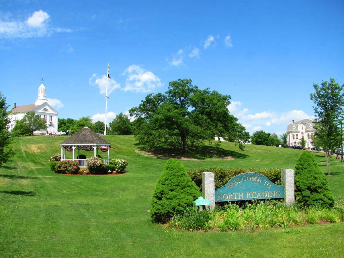 Green park with a sign that says "Welcome to North Reading," flowers, a gazebo with patriotic decorations, a large tree, two church buildings, and a clear blue sky.