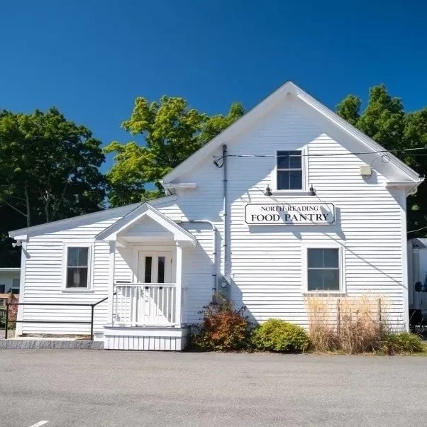 White building with a sign for North Reading Food Pantry, surrounded by trees and bushes, under a blue sky.