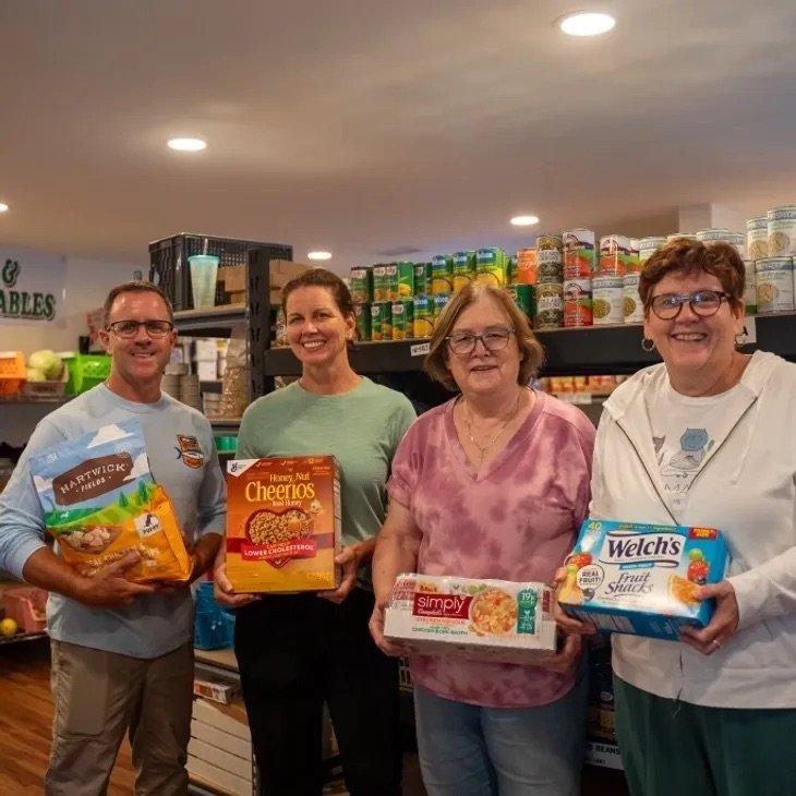 Four people smiling inside a grocery store, each holding a box of food or snack items.