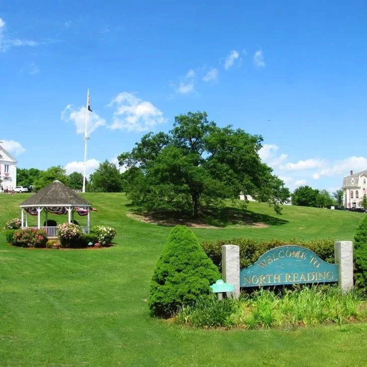 Welcoming park area with a sign that says 'Welcome to North Reading', a large green tree, a smaller bush, and a gazebo decorated with American flags, under a clear blue sky.