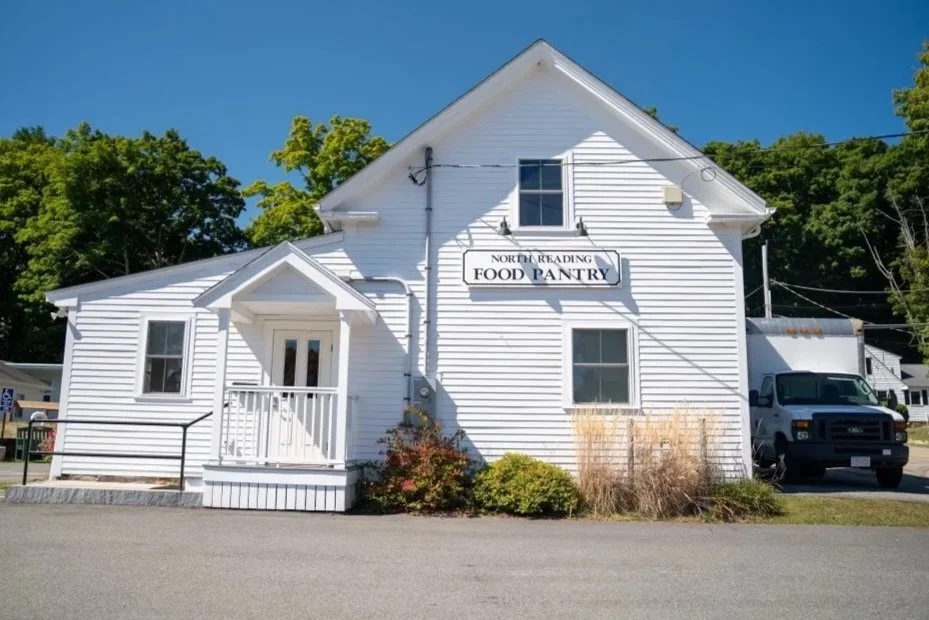 White two-story building with a sign that reads 'North Reading Food Pantry,' a small porch with a white railing, and a black pickup truck parked beside it, surrounded by green trees and a clear blue sky.