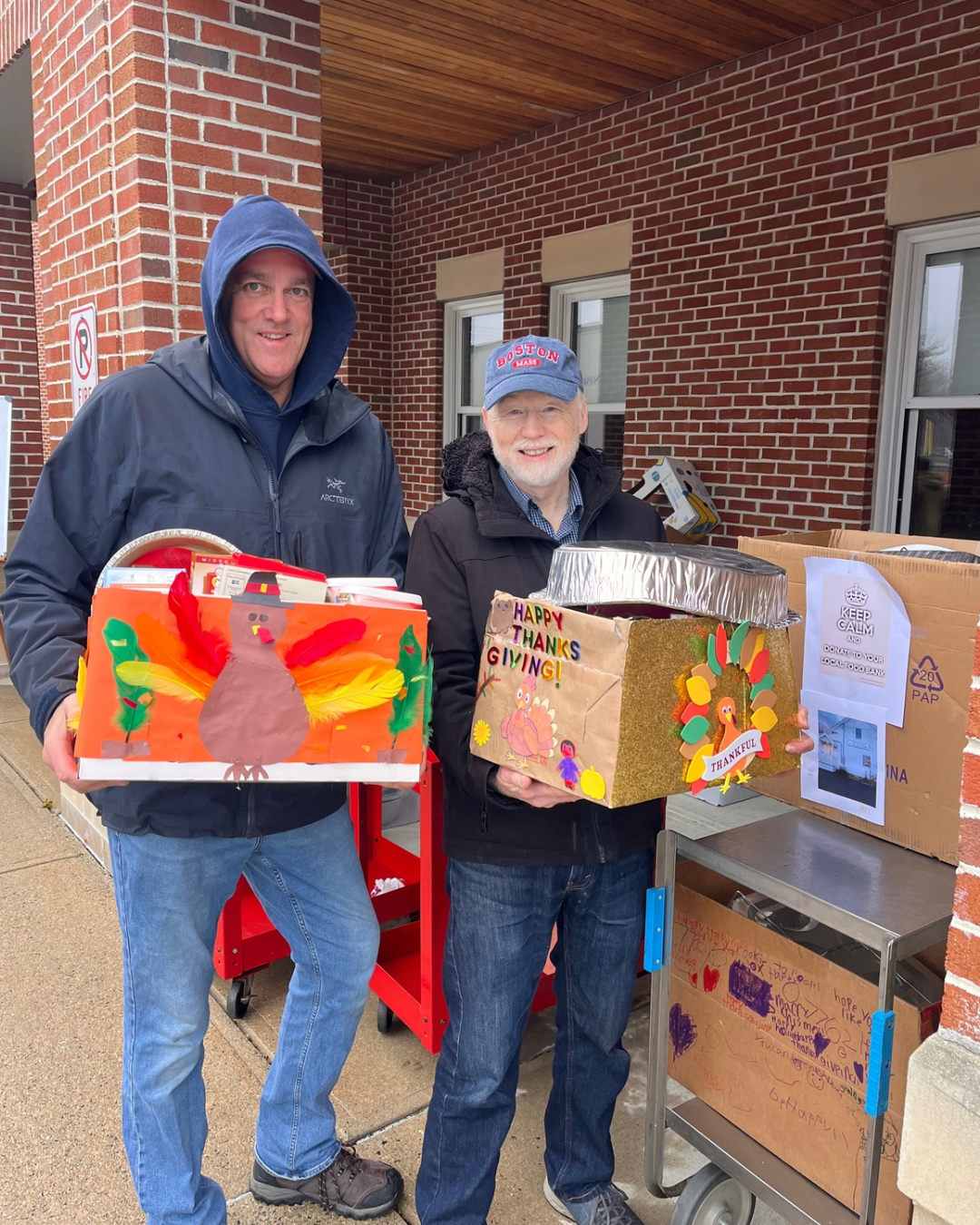 Two men holding decorated boxes stand outside near a brick building. One wears a black jacket and the other a cap with 'Boston' written on it. They are participating in a donation or community event.