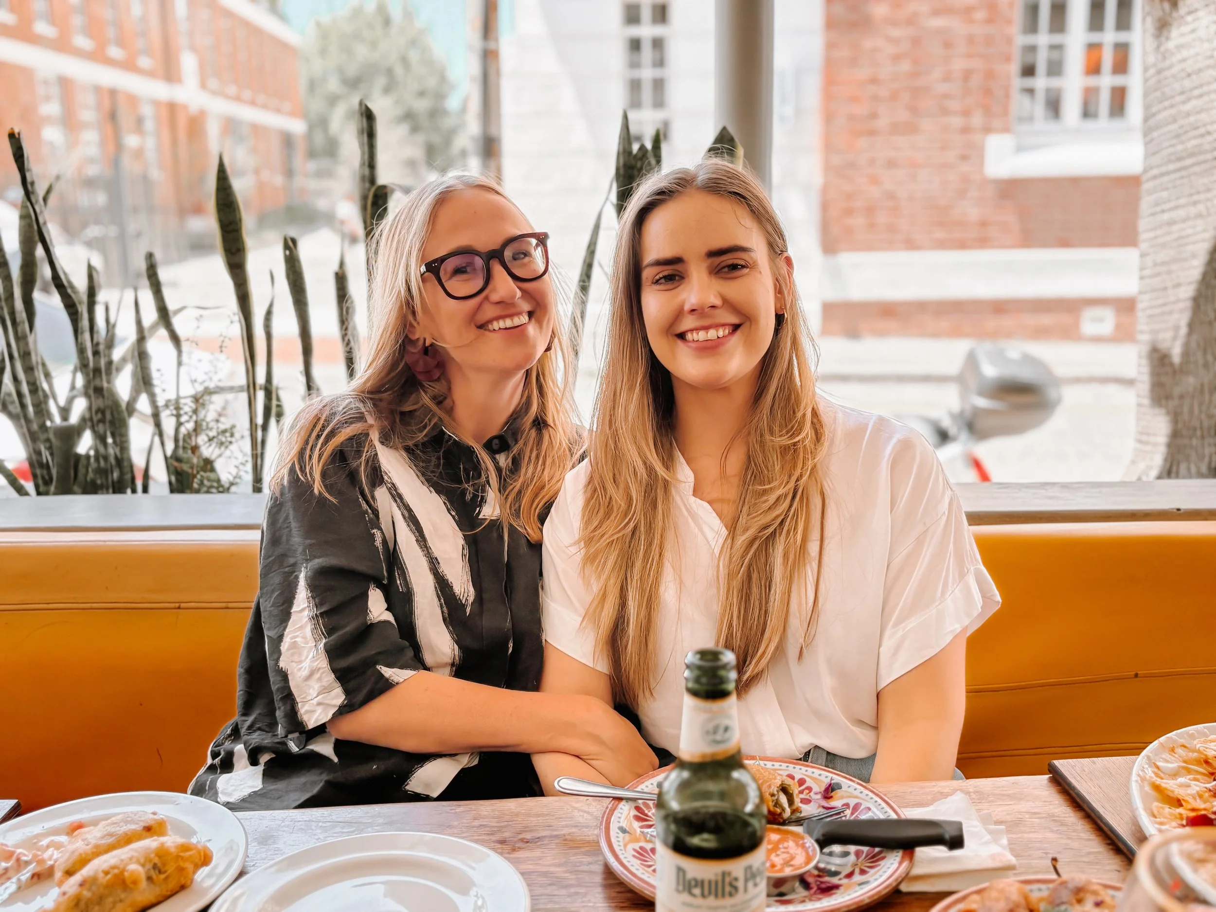 Two women sitting together at a dining table inside a restaurant, smiling at the camera. There are pizza and other dishes on the table, along with a beer bottle in front of the woman on the right. The background shows windows with a view of brick buildings and trees outside.