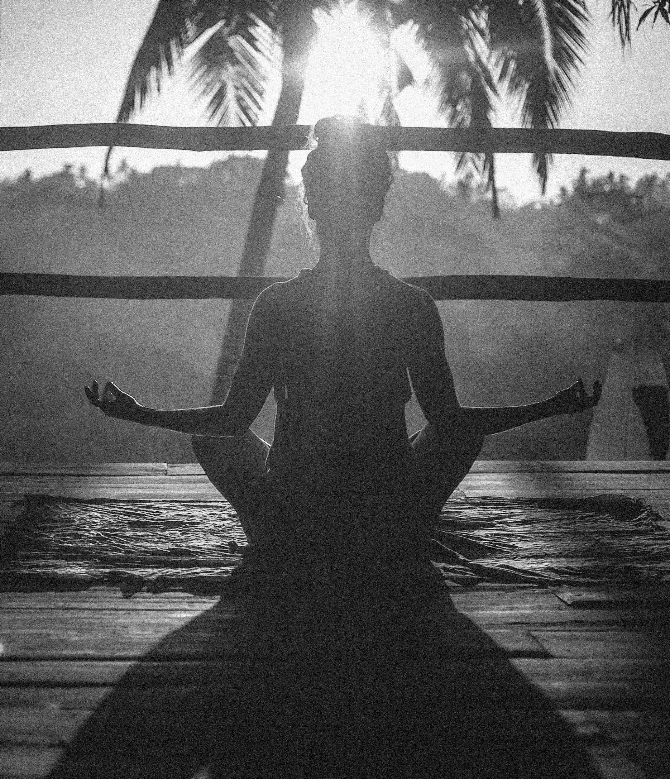 Silhouette of a person sitting in a lotus pose on a wooden surface, facing the ocean with palm trees in the background, during sunset