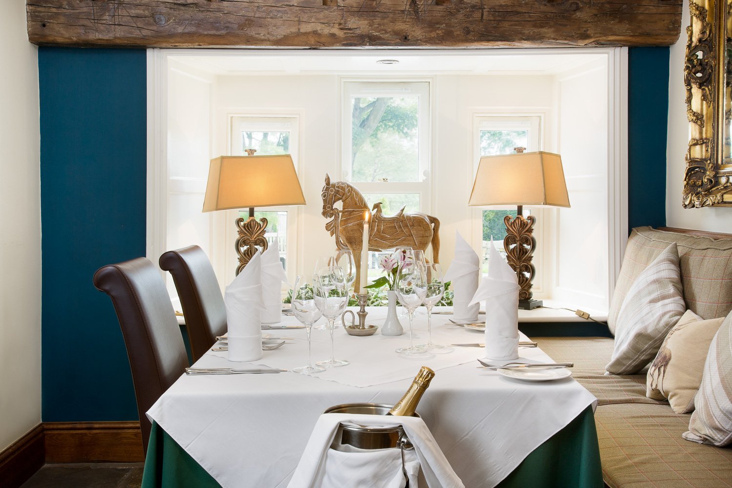 A dining room with a table set with wine glasses, napkins, and a centerpiece with a wooden horse sculpture and pink flowers. Two table lamps with ornate bases are on side tables by a window overlooking a green outdoor area. There are upholstered chairs and a built-in bench with plaid cushions along the window wall.