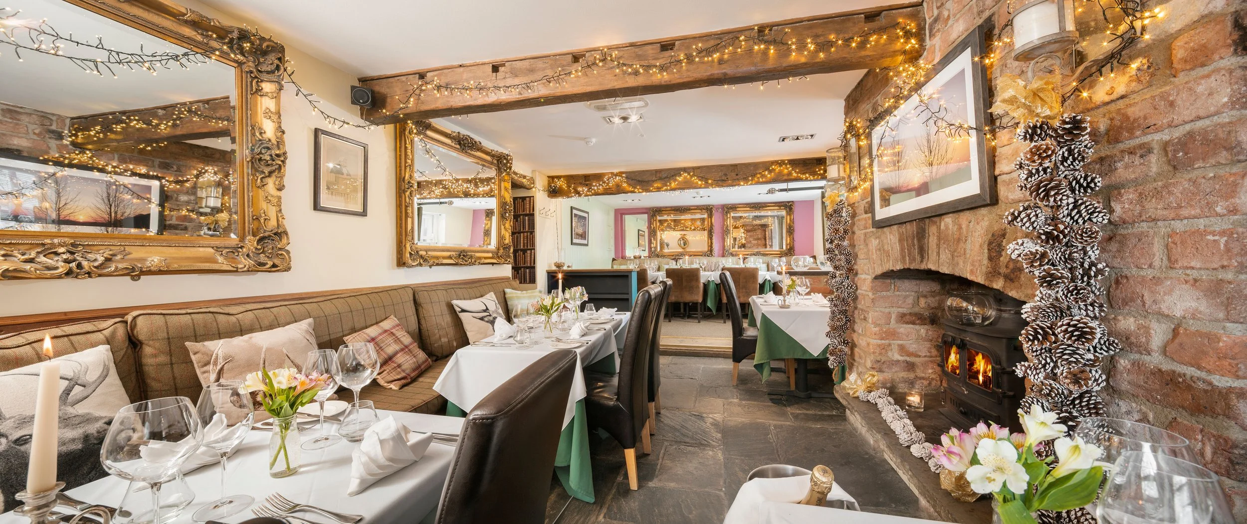 Cozy restaurant decorated with string lights, gold-framed mirrors, and holiday pinecone garlands, with tables set for dining and a brick fireplace.