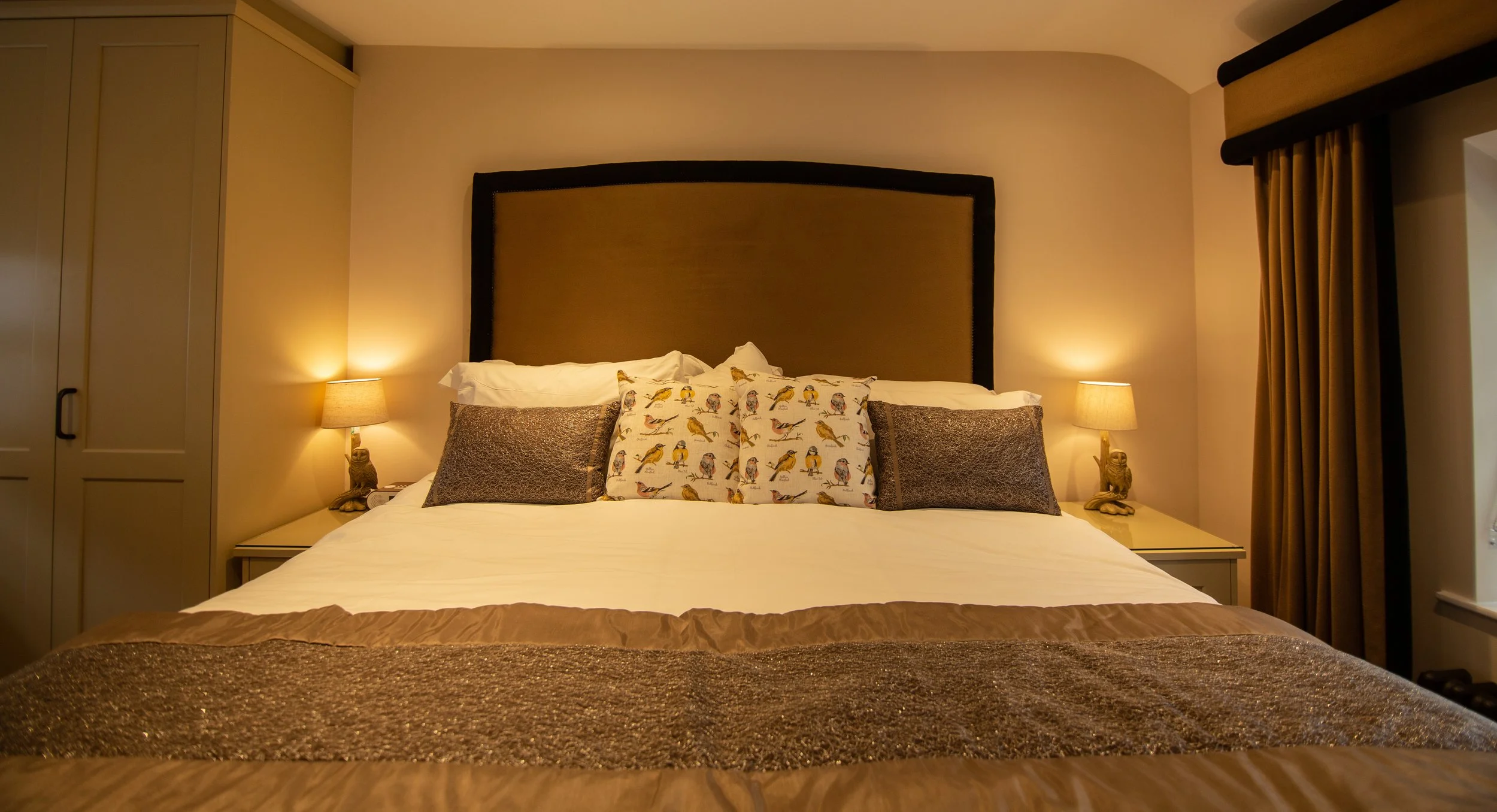 A neatly made bed with multiple pillows, including two with bird patterns and two brown textured pillows, in a warmly lit hotel room with beige walls, side tables, lamps, and curtains.